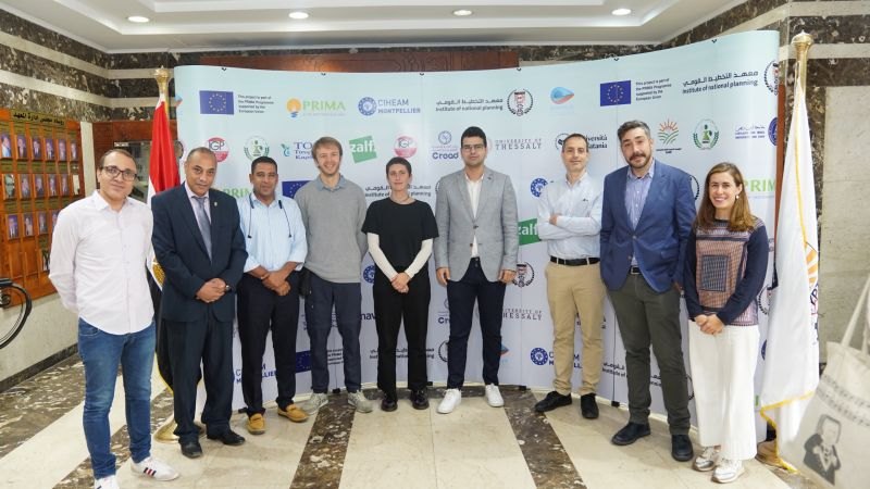 Nine meeting participants in front of the DIONYSUS partner logo wall, which includes the University of Thessaly